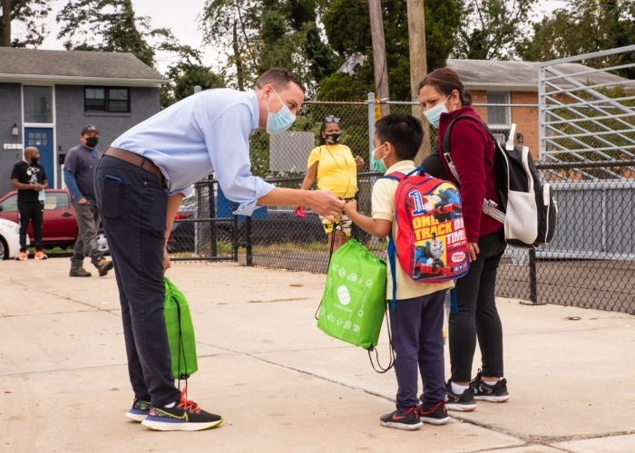 Masked DCSEU staff member passing out lighting kit bag to a masked elementary school student and their parent.