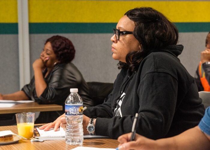 A workforce development extern sitting at a desk taking notes next to her classmates and listening to an instructor.