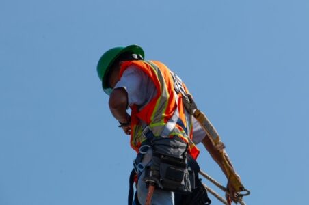 The back of a solar installer on a roof with the sky as a backdrop, adjusting their safety gear.