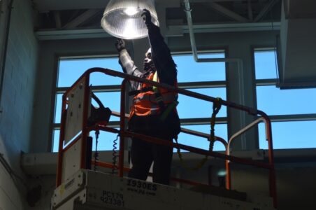 Installer on a scissor lift reaching up to finish installing an LED High-bay fixture.
