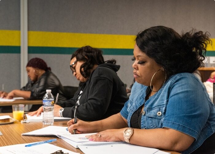 A row of workforce development program externs sitting at a desk taking notes in a classroom and office setting.