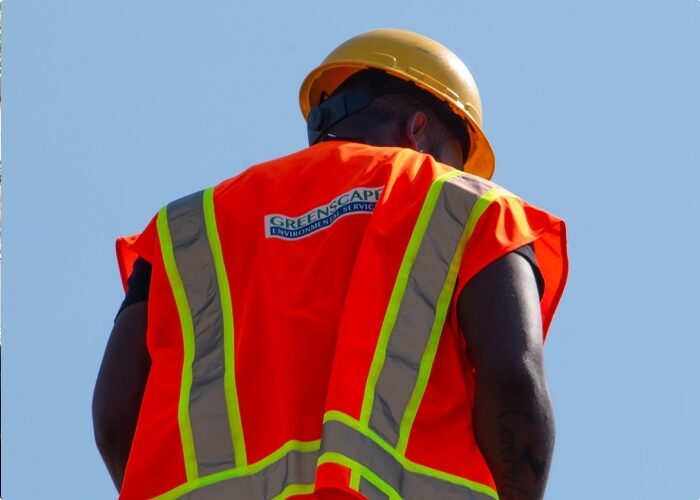 Closeup of the back of a construction worker with an orange vest and yellow hardhat against the blue background of the sky.