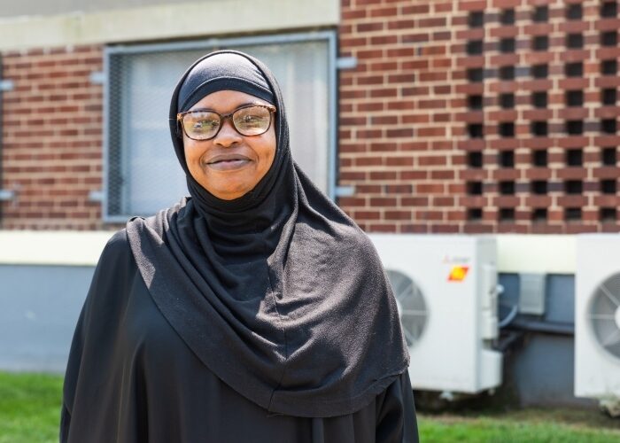 A woman in a black jilbab smiling in front of an affordable apartment complex with heat pump compressors visible.