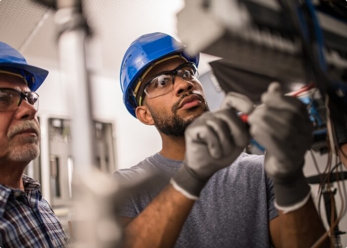 A worker in a hardhat, safety glasses, and gloves working on a commercial electrical panel while being supervised by another worker.
