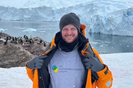 DCSEU staff member, Doug Abbott smiling in the arctic with his jacket open to show the DCSEU logo on his shirt. He is standing in front of an oceanside rock formation with penguins on it.