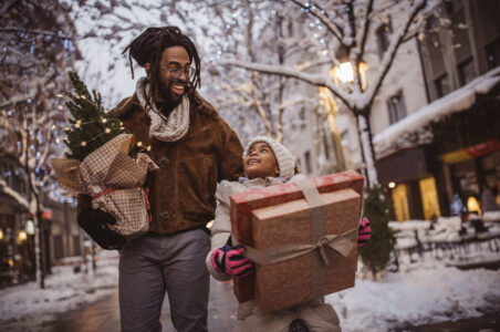A father and child walking down a snowy urban street with holiday gifts and a small holiday tree in hand while they smile at each other.