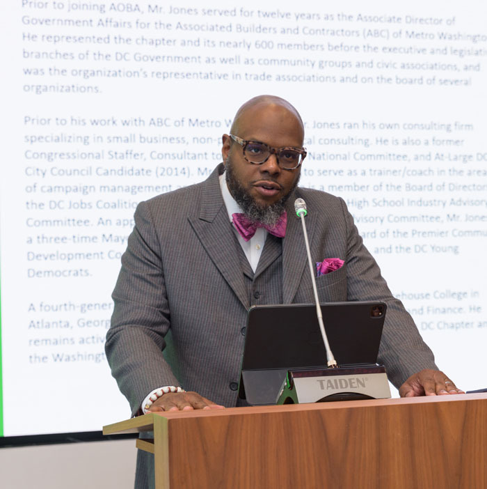 Eric J. Jones, AOBA VP of Government Affairs, at a podium in front of a screen with his bio on it providing the commencement address to graduating externs.