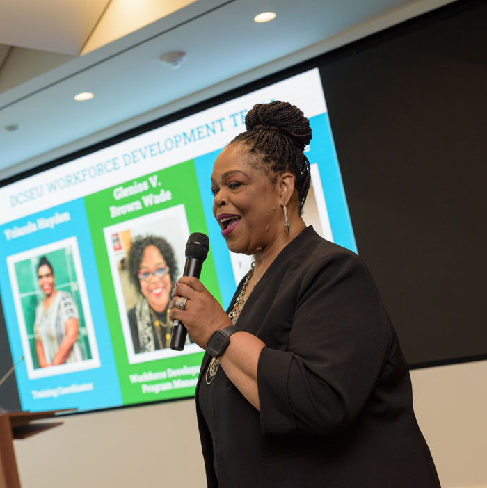Workforce Development Program Manager, Gleniss V. Brown Wade, providing remarks to externs and graduation attendees. She is standing in front a screen with images of the program operations team.