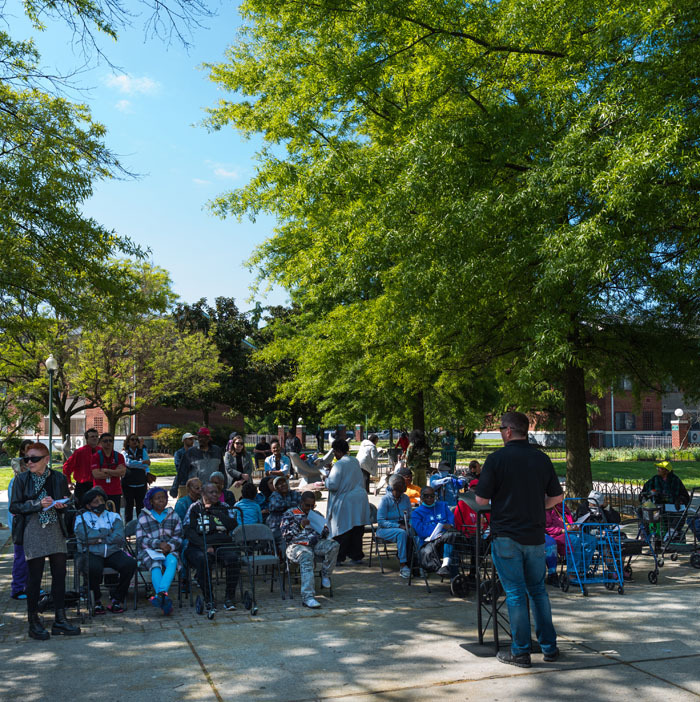 Silas Naecker, President, John G. Webster, providing remarks to a large, seated group of Parkside residents in front of their community center in the communal courtyard.