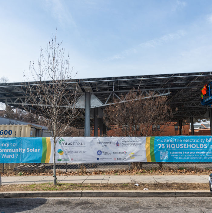 The Sargent Memorial Presbyterian Church's community solar canopy from street level with a banner in front of it on a parking lot fence.
