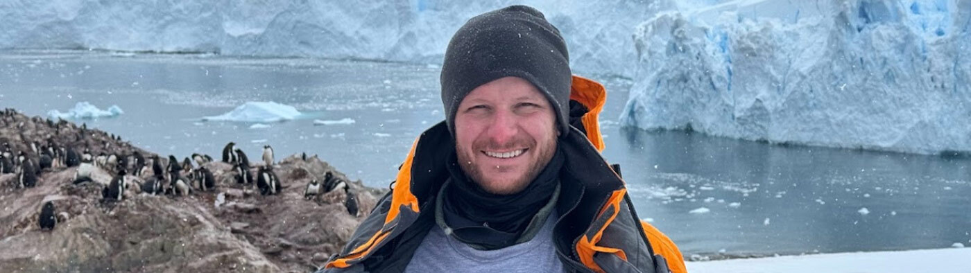 DCSEU staff member, Doug Abbott smiling in the arctic with his jacket open to show the DCSEU logo on his shirt. He is standing in front of an oceanside rock formation with penguins on it.