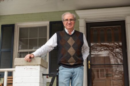 Bob, an older man with gray hair, glasses, and a navy and brown argyle sweater vest with white dress shirt underneath standing on his porch in Ward 3, DC. He is holding on to the porch banister with his right hand and has his left hand in his back pocket.