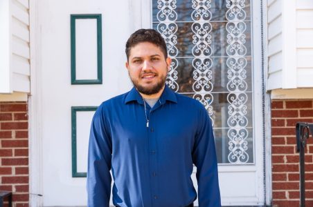 Victor posing in front of his home's front door with a blue oxford shirt, smiling.