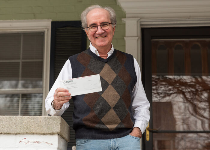 Bob, an older man with gray hair, glasses, and a navy and brown argyle sweater vest with white dress shirt underneath standing on his porch in Ward 3, DC. He is holding his DCSEU rebate check in his right hand, showing it toward the camera and smiling.