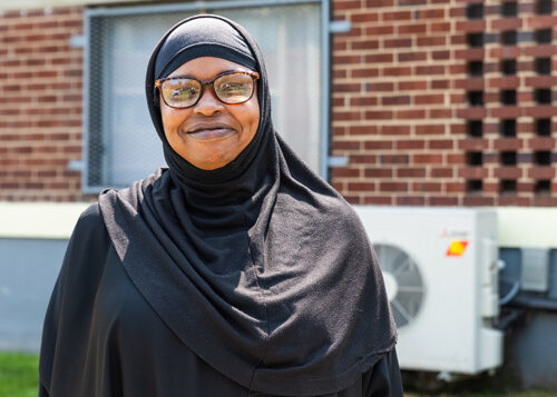 Chandra Williams, Paradise at Parkside resident, in a black jilbab smiling in front of an affordable apartment complex with heat pump compressors visible.