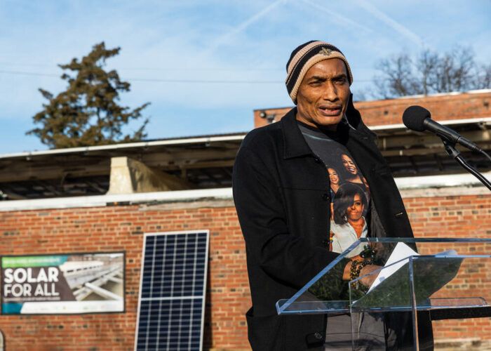 Fairfax Village Community Association President, Lem Walker, standing at a podium providing remarks in front of one of the villages many CREF solar systems.
