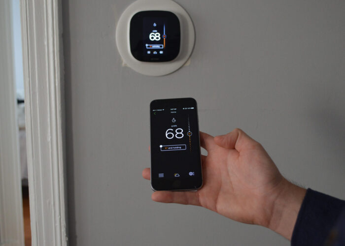 hand holding a smart phone in front of a smart thermostat mounted to a wall with the app for the thermostat open on the phone showing the temperature set to 68 degrees Fahrenheit.