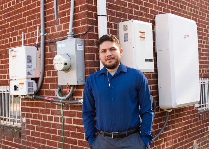 Ward 7 resident, Victor Ramos, in a blue oxford shirt standing in front of his home's new solar inverter and battery system provided in a solar battery pilot for DC's Solar for All Program.