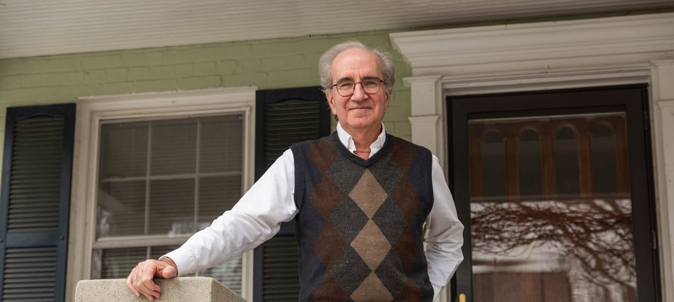 Bob, an older man with gray hair, glasses, and a navy and brown argyle sweater vest with white dress shirt underneath standing on his porch in Ward 3, DC. He is holding on to the porch banister with his right hand and has his left hand in his back pocket.