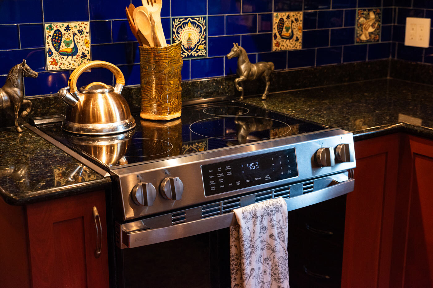Silver and black induction range with a copper tea pot and utensil holder sitting on the stove stop.  The stove is nestled between granite countertops and a blue title backsplash.