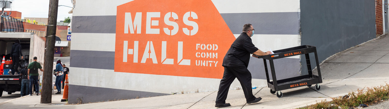 Culinary worker pushing a cart in front of the Mess Hall building mural.