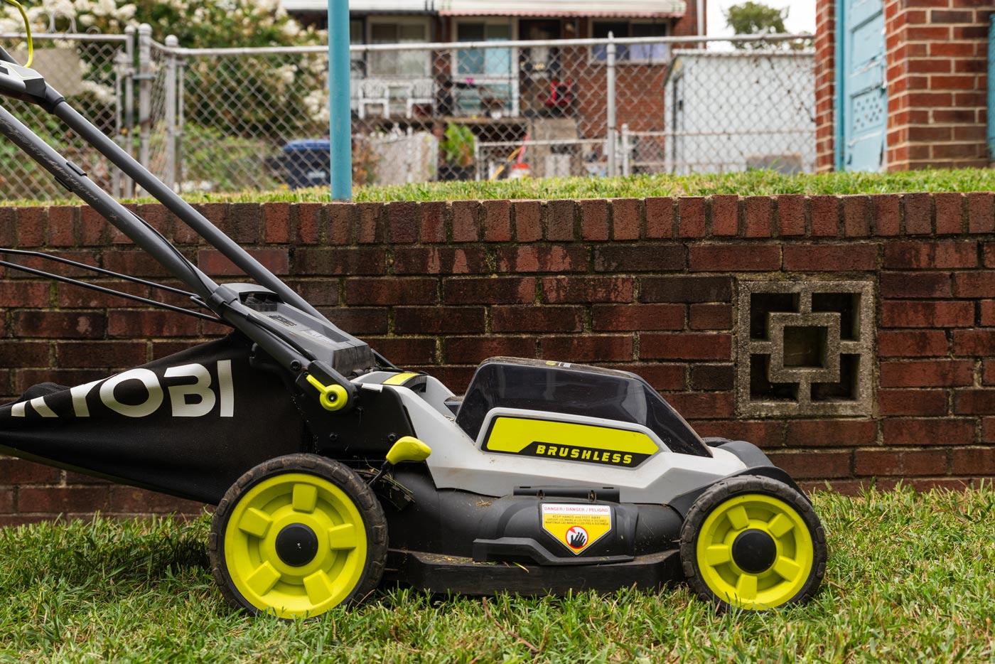 Black electric lawn mower with lime green trim in a small DC backyard on freshly cut grass with a brick wall and alleyway visible in the background.