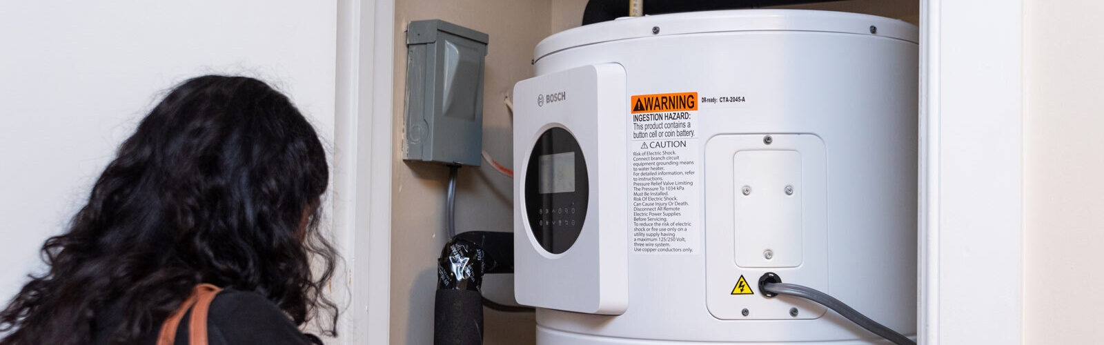 A person looking at a white Bosch heat pump water heater within the utility closet of a unit within Victory Square Senior Apartments.