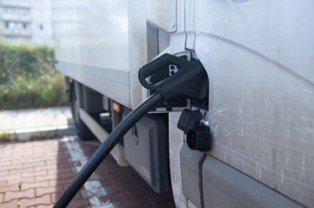 Close up picture of a black EV charger handle plugged in to a white zero-emissions freight truck. The side of the truck has moderate wear and tear from use.