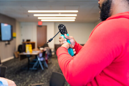 Young and bearded African American man in a red hoodie in a training class. He is learning how to use wire cutters to strip a wire for solar panel applications.