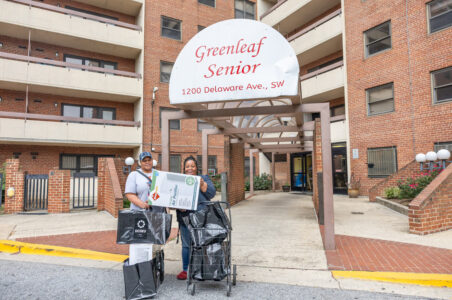 Two African-American residents of Greenleaf Senior public housing standing at the front entrance of the building hold air purifiers from their new free energy conservation kits.