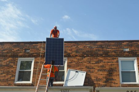 Solar installer in orange shirt on top of DC row home porch overhang passes a solar panel up to another installer on the roof on sunny day.