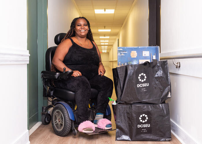 African-American Greenleaf Senior resident that is wheelchair bound in front of her apartment posing and smiling with her new DCSEU-branded free energy conservation kit that includes two air purifiers, an advanced power strip, and Wi-Fi-enabled smart plugs.