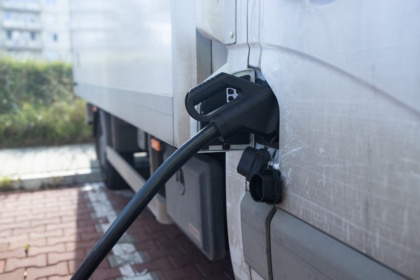 Close up picture of a black EV charger handle plugged in to a white zero-emissions freight truck. The side of the truck has moderate wear and tear from use.