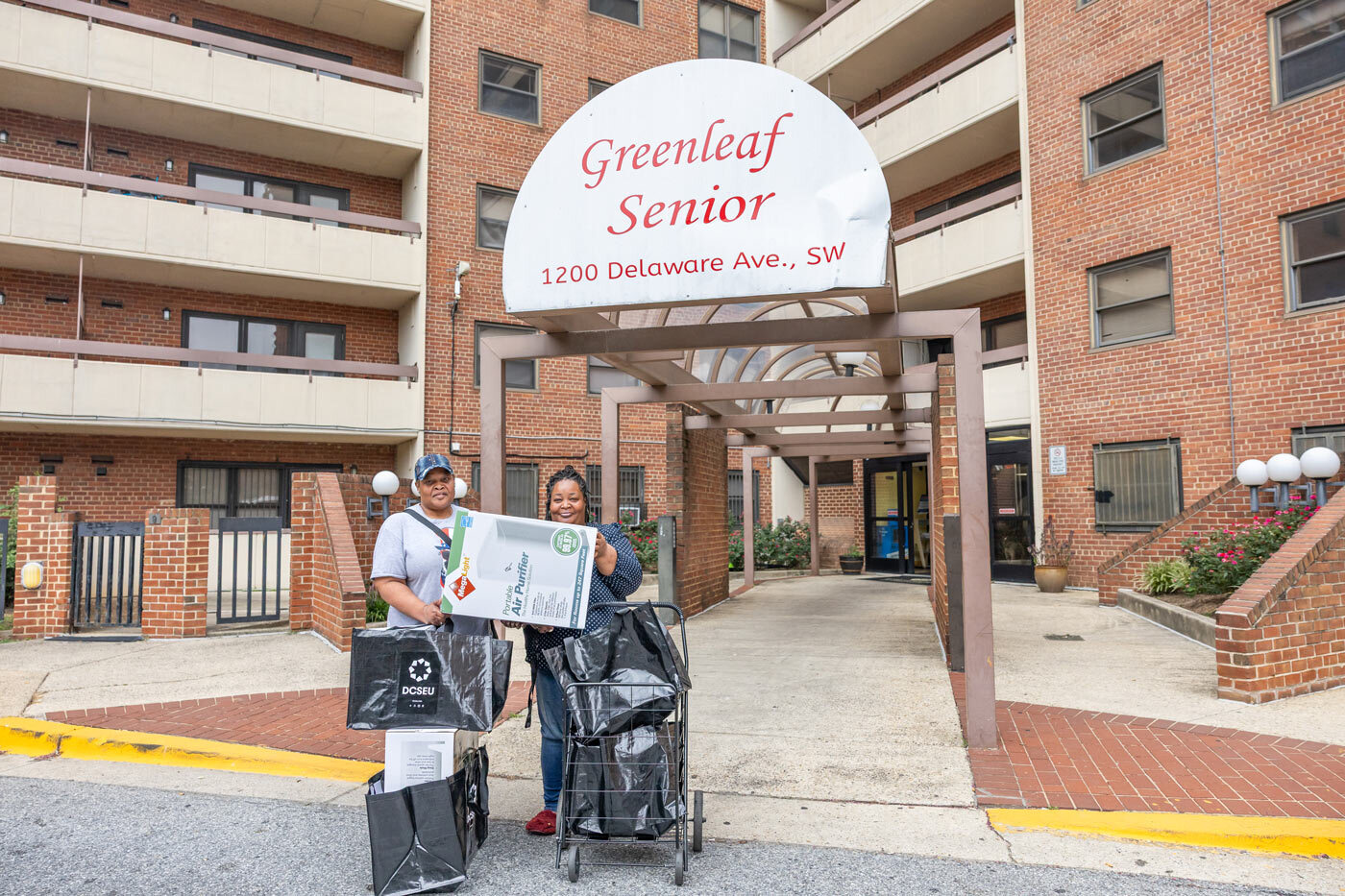 Two African-American residents of Greenleaf Senior public housing standing at the front entrance of the building hold air purifiers from their new free energy conservation kits.