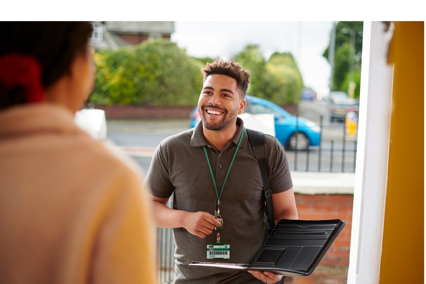 A young, African American, masc-presenting sales person smiling at the door of a single-family home.  The resident of the home stands in the doorway listening as the sales person shows his badge and portfolio.