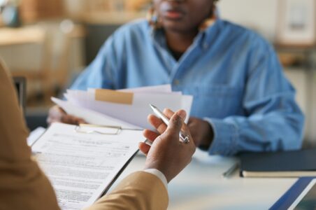 Close up of the hands of a tax accountant reviewing tax returns with a client sitting across from them. The accountant is pointing to parts of the document while the client is holding additional forms and listening intently.