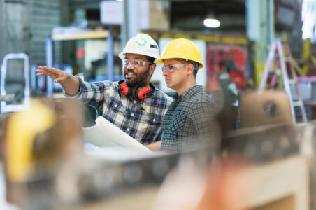 Two workers in hardhats reading blueprints in a warehouse and point to something in the distance.