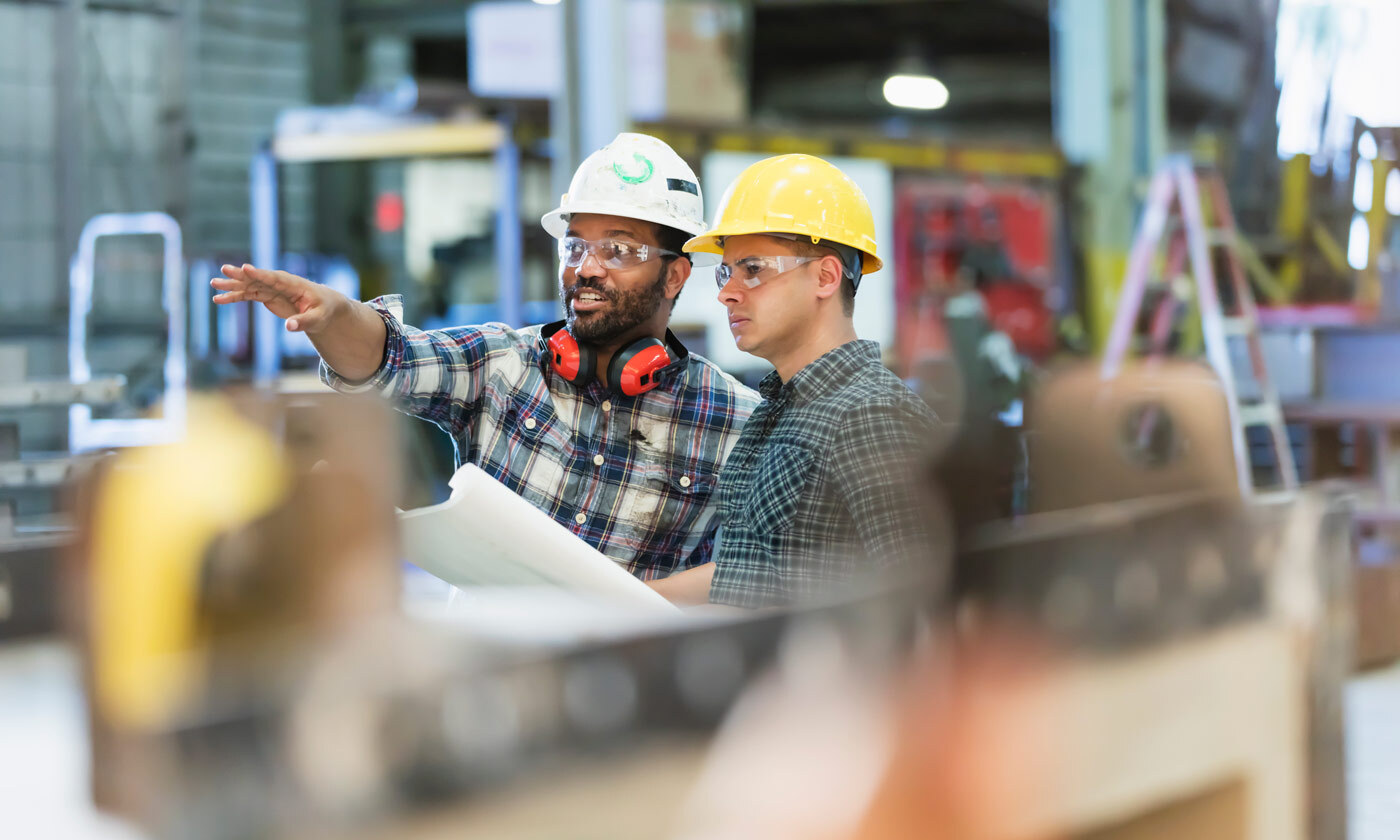 Two workers in hardhats reading blueprints in a warehouse and point to something in the distance.