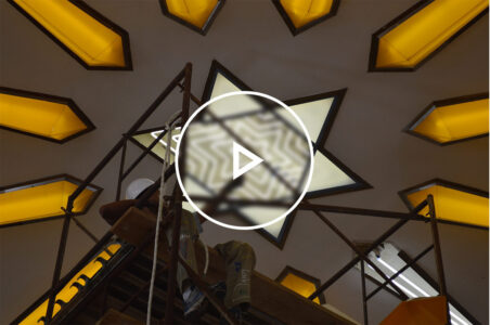 Electrical worker sitting on scaffolding below a neon star of david light fixture in the process of changing out the tubes with a play button graphic over the image.