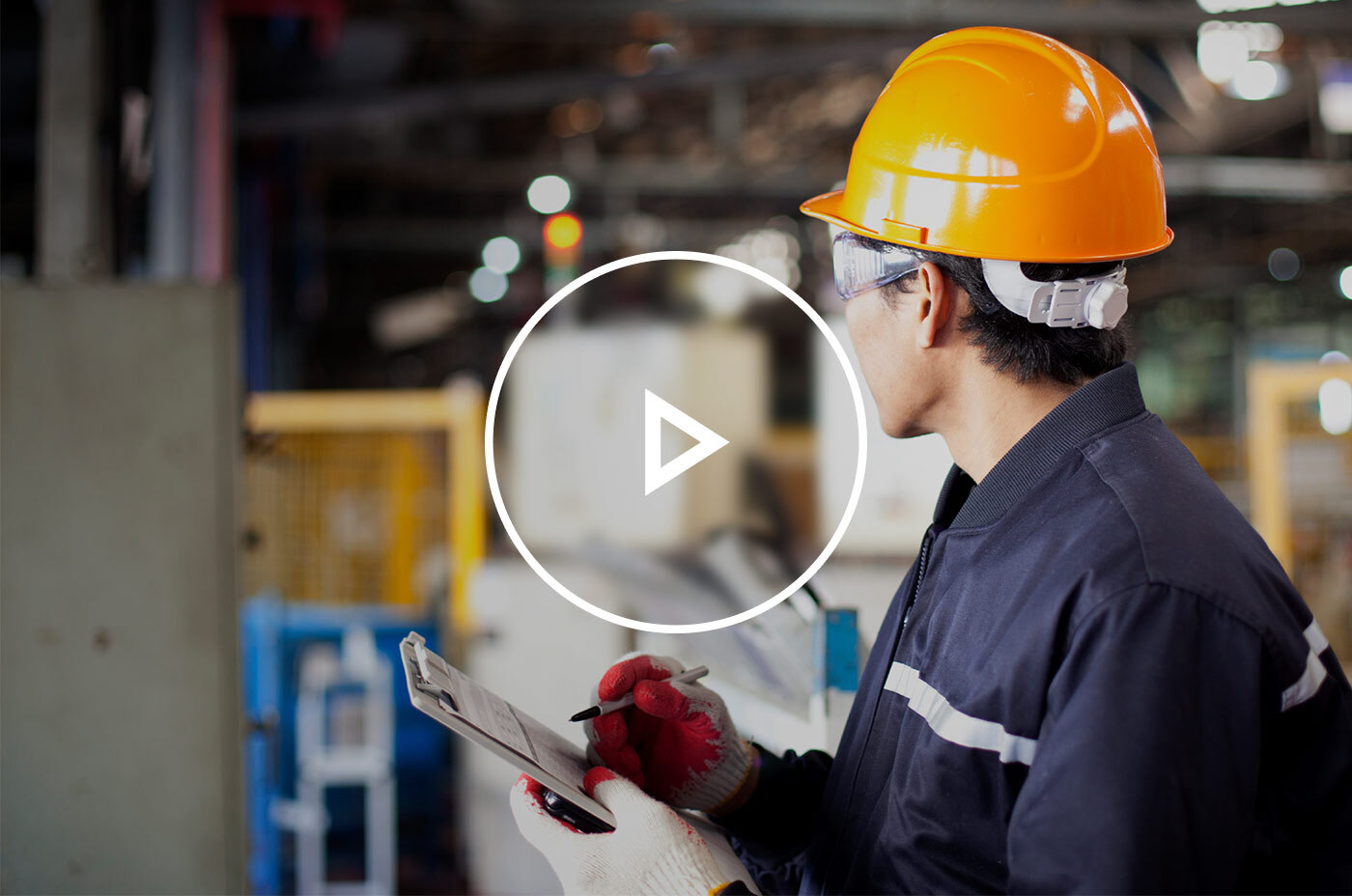 Contractor with a yellow hard hat on in a warehouse looking away from the camera as he inspects and writes down notes on a clipboard.