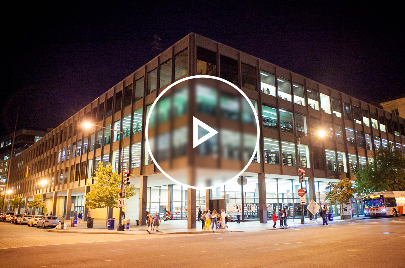 The Martin Luther King Jr. Library building facade at nighttime with streetlights on and different lights on within the building. People are walking in front of the building as well.