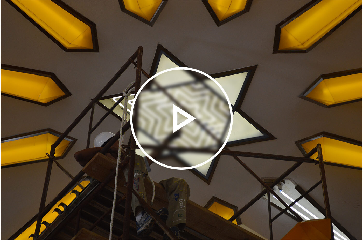 Electrical worker sitting on scaffolding below a neon star of david light fixture in the process of changing out the tubes with a play button graphic over the image.