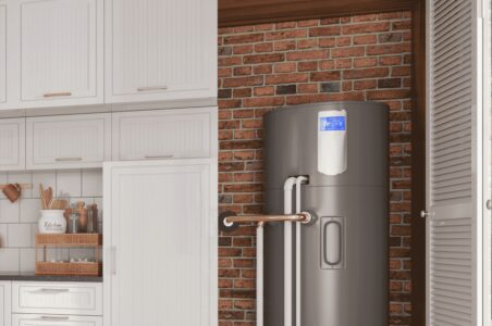View of a kitchen with white cabinets and accents with the utility closet open. Inside the closet against a brick wall is a grey heat pump water heater with pipes entering the tank along with the floors and walls of the home.