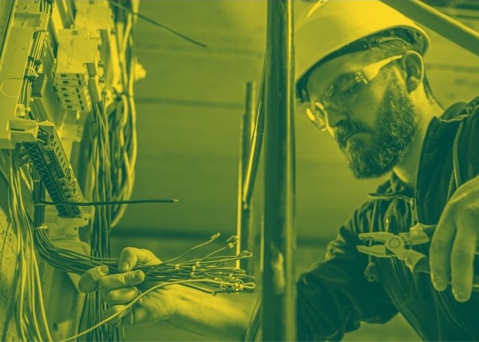 Green duotoned image of an electrician wearing protective eyewear and helmet with plyers in his hands working on installing the circuits within an electrical panel.
