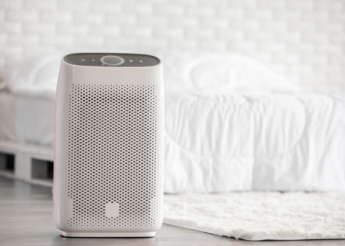 An air purifier at the foot of a white bed.